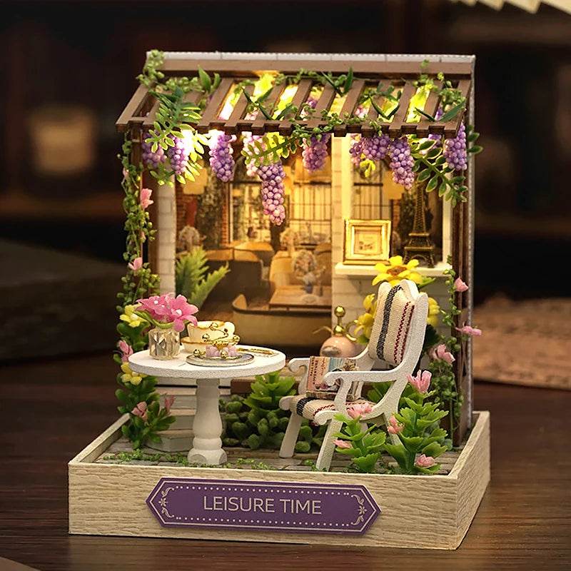 Miniature DIY dollhouse garden with white table, pink flowers, rocking chair, and hanging purple wisteria.
