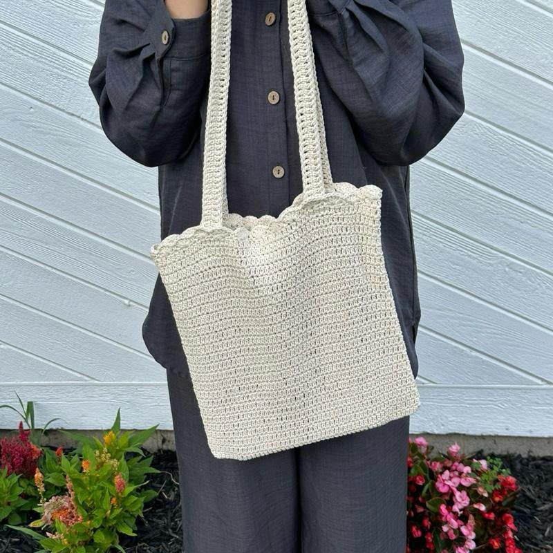 Beige handmade crochet tote bag with scalloped top edge and long handles worn over black outfit.