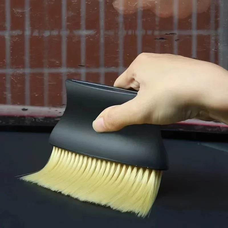 Hand holding black ergonomic car interior dust brush with soft yellow bristles cleaning dashboard grooves.