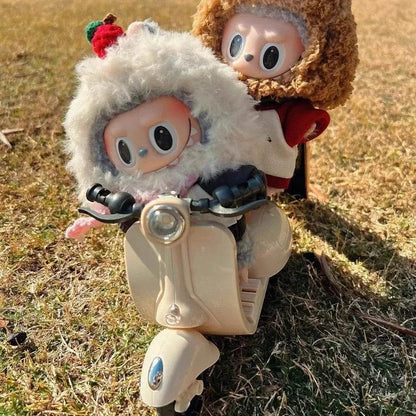 Beige Labubu doll wearing fluffy hood riding mini electric motorcycle on grass with brown-haired doll passenger.