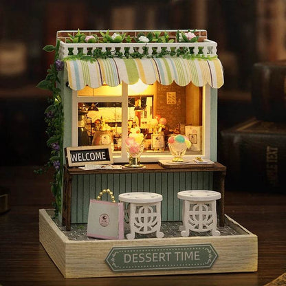 Miniature flower house dollhouse with striped awning, warm interior lighting, two white stools, and "Dessert Time" sign on.