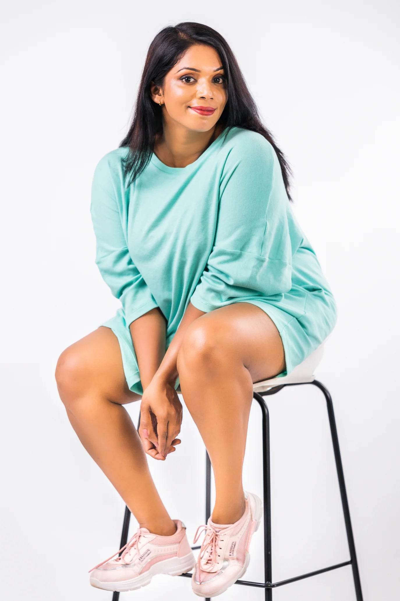 Relaxed fit mint green long sleeve t-shirt worn with pink sneakers, seated on black stool.