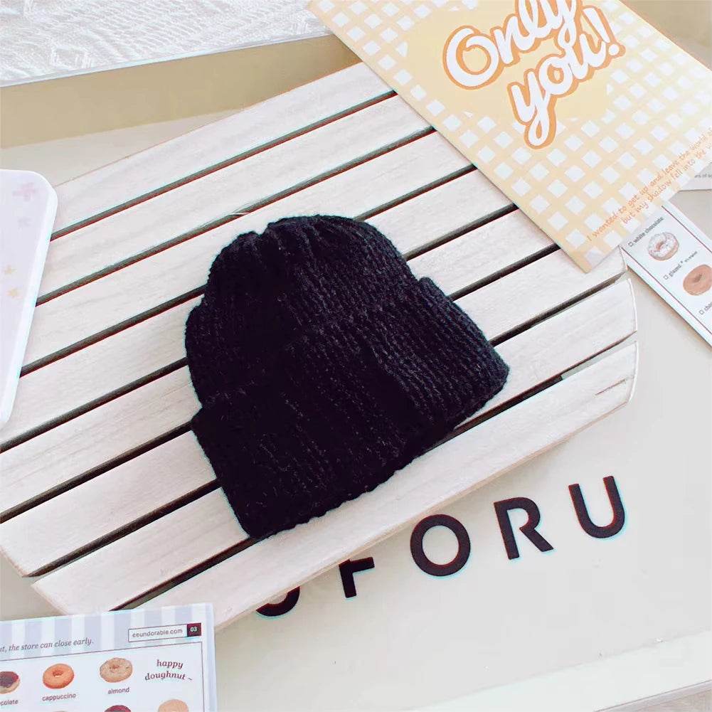 Black knitted beanie hat for 17cm dolls on white slatted surface with decorative cards nearby.