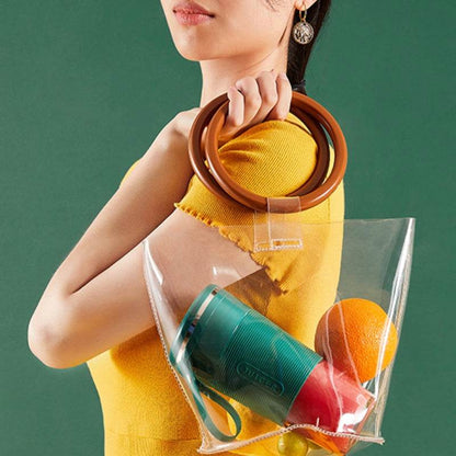 Transparent tote bag with brown wooden handles holding a green and pink portable blender and an orange.
