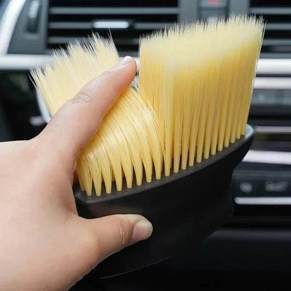 Hand holding black car interior cleaning brush with dense soft cream bristles near vehicle dashboard controls.