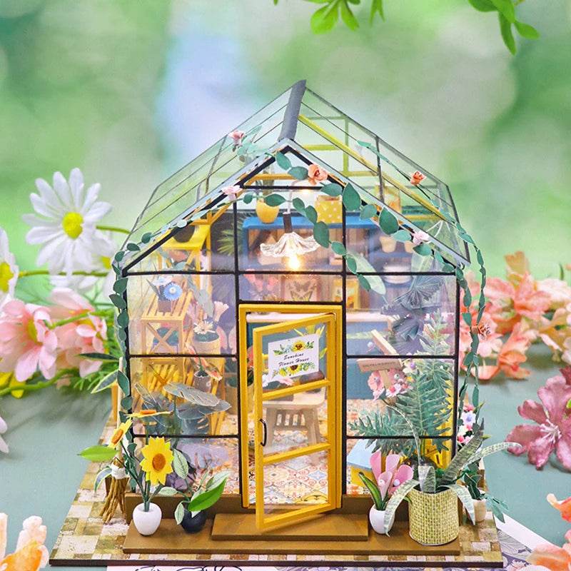 Transparent miniature flower house with yellow door, detailed interior furniture, and potted plants outside.